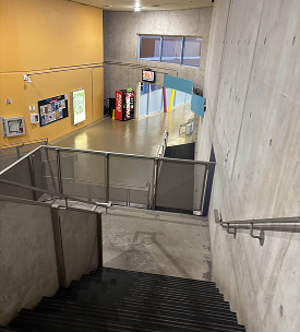 Top of the main staircase of J wing, outlooking onto the main entrance/lobby area. There is the bulletin board, a vending machine, and plenty of seats. There is an arch and it is a very open area leading out into a hallway with more classes.